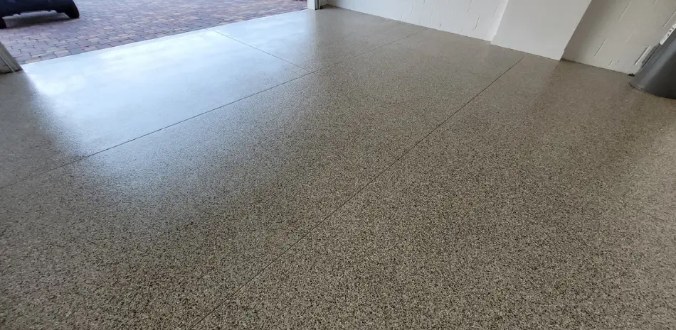 A garage with a newly installed epoxy flake floor, showing a speckled pattern of black, grey, and white. Sunlight reflects off the smooth, glossy surface near the garage opening.